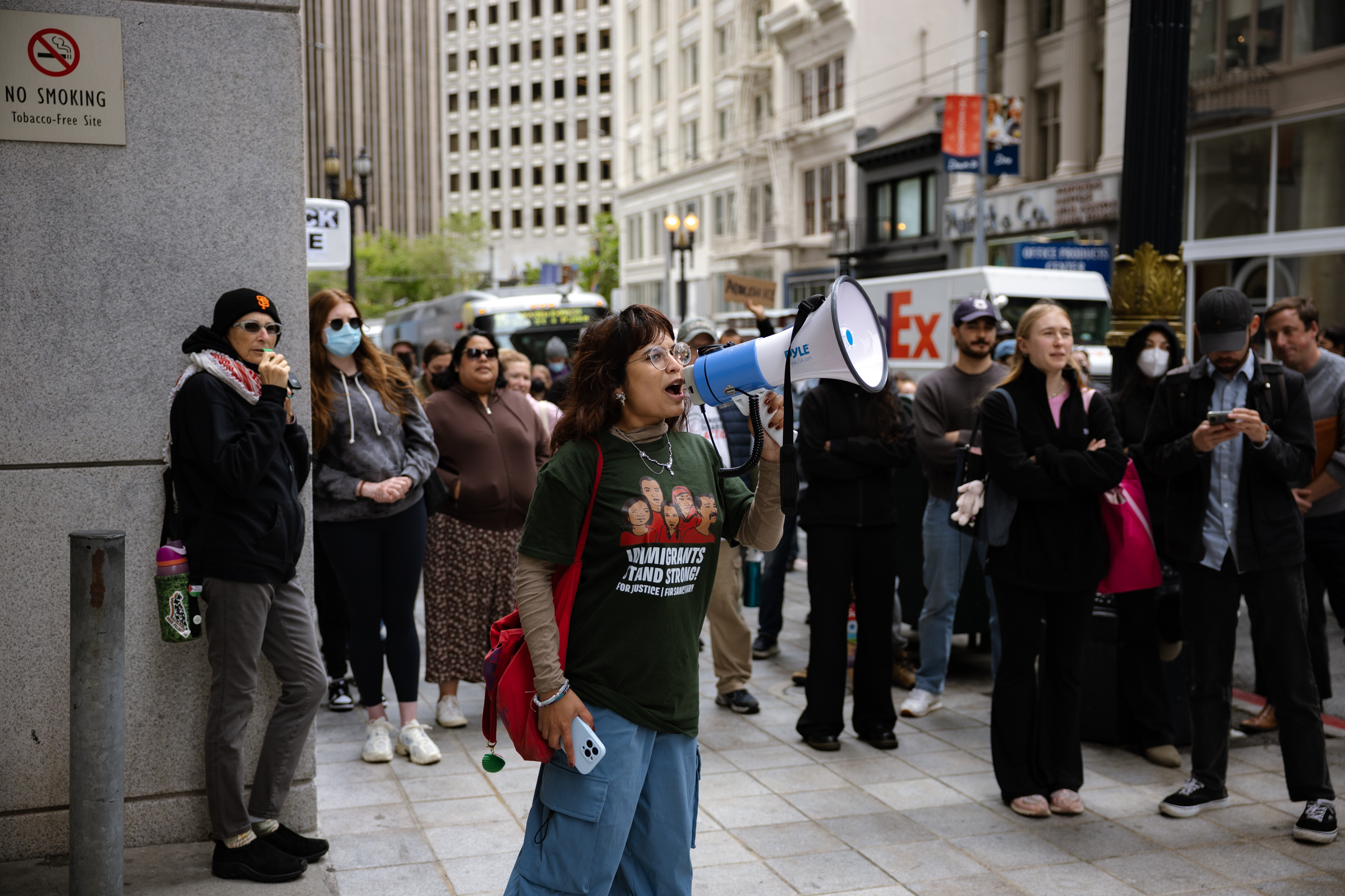 A person uses a megaphone, leading a protest or rally. A diverse group stands in the background, some with masks, observing in a city street setting.